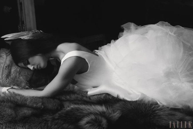 Model Dillon Collins lying on a fur rug wearing a voluminous white tulle wedding gown and feather headpiece in black and white.