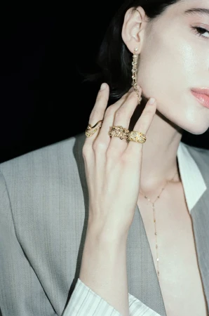 Close-up of model Erika Barletta wearing textured gold rings, a drop earring, and a grey tailored blazer against a dark background.
