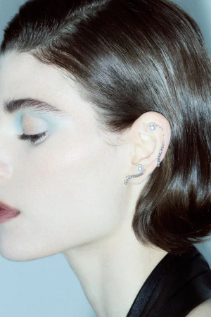 Close-up profile of a woman wearing silver and pearl ear cuffs with soft blue eyeshadow and short dark hair against a pale backdrop.