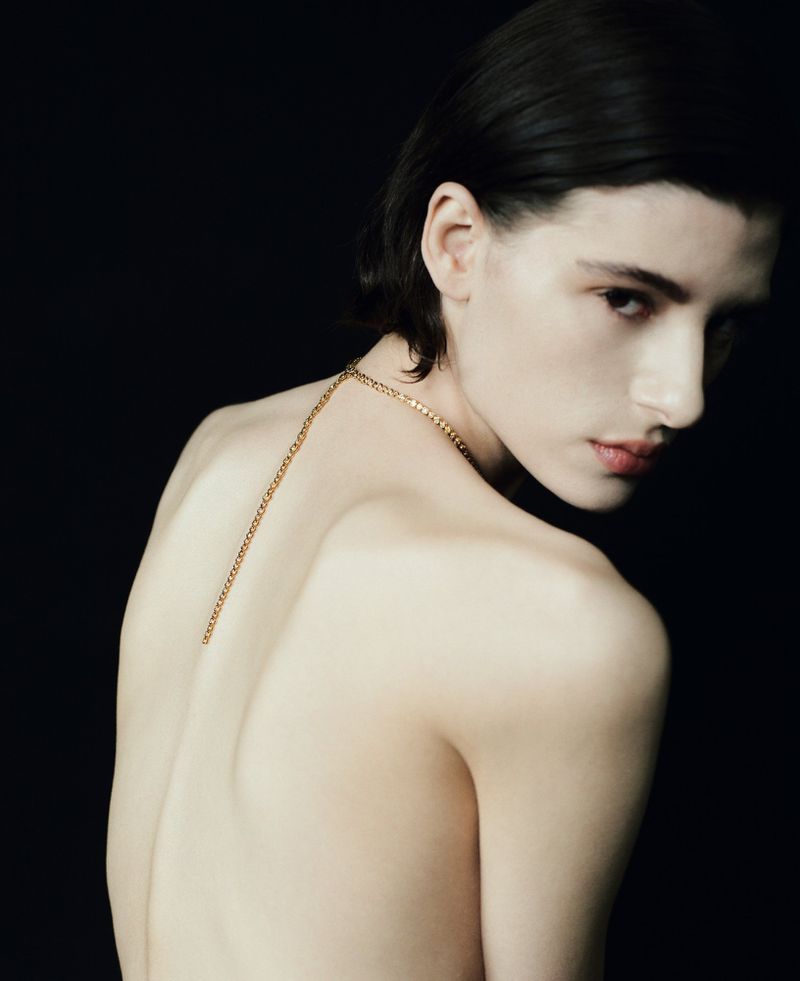 Model Erika Barletta looking over her shoulder with a gold chain draped down her bare back against a solid black background.
