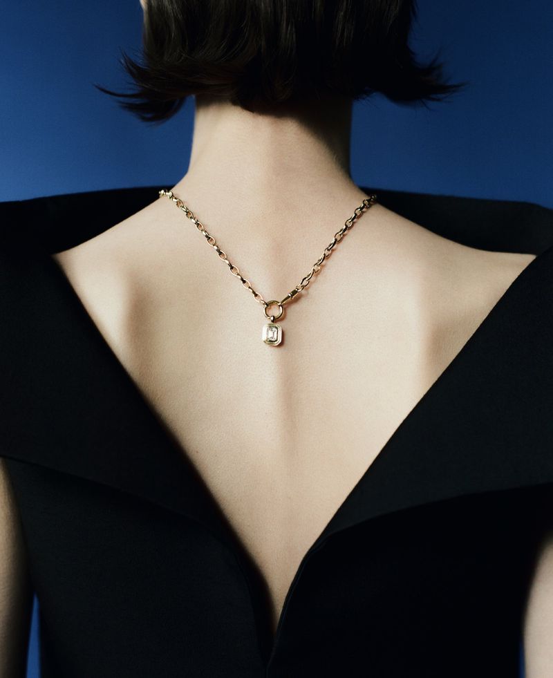 Model Erika Barletta seen from behind wearing a gold pendant necklace against a blue studio background.