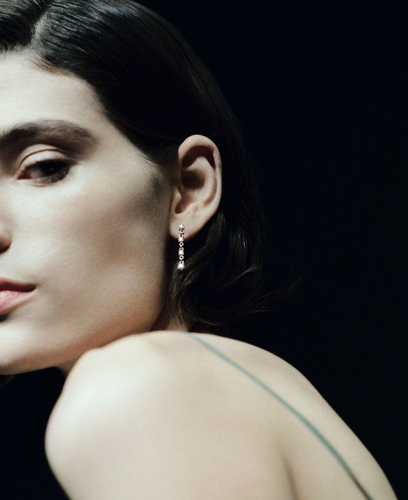 Close-up profile of model Erika Barletta wearing a delicate diamond drop earring against a solid black background.