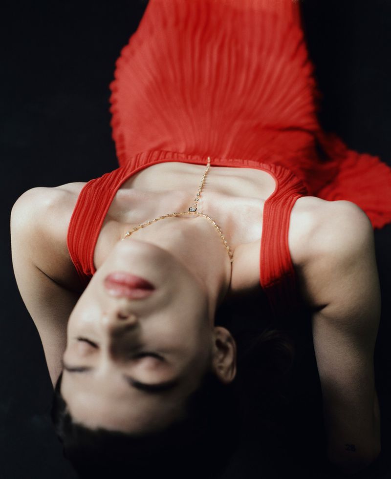 Model Erika Barletta leaning back in a pleated red dress and delicate gold pendant necklace against a dark background.