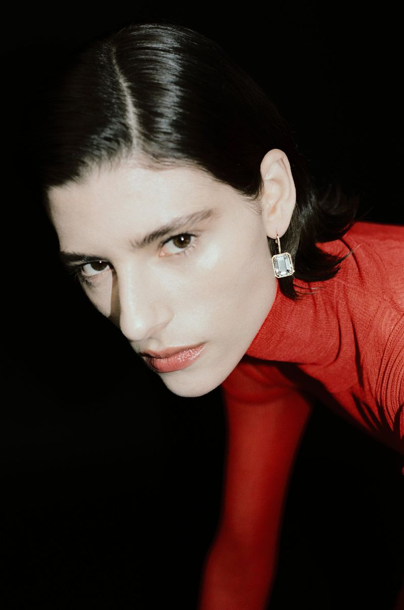 Model wearing a sparkling rectangular crystal drop earring and a fitted red sheer top against a solid black background.