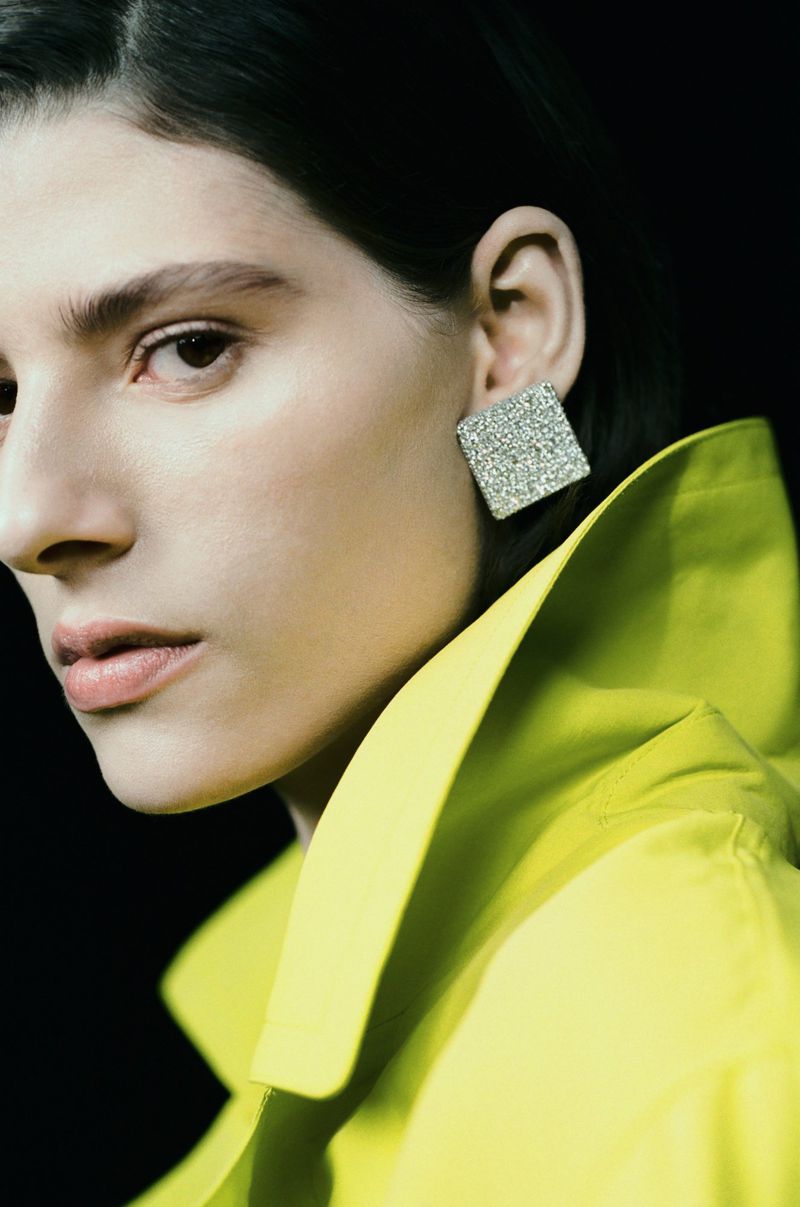 Close-up portrait of a woman wearing a lime green collared jacket and a large square silver earring against a dark background.