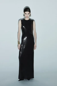 Model Erika Barletta in a full-length sleeveless black sequin gown with a gold cuff bracelet against a light grey studio background.