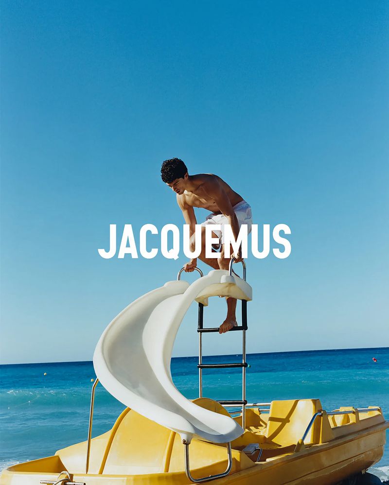 A man in white swim shorts stands on a curved slide attached to a yellow paddle boat in turquoise ocean water with Jacquemus branding.