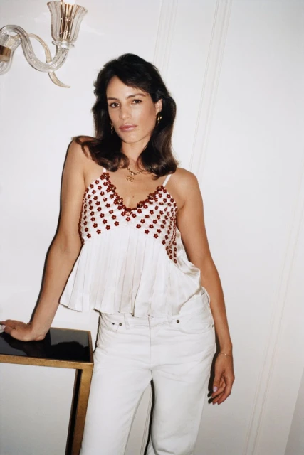 Portrait of a brunette woman indoors wearing a white tank top, white pants, and gold jewelry leaning against a black and gold table.