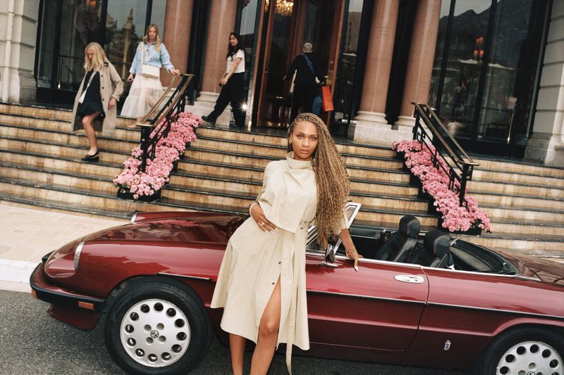 Woman in tan dress poses next to a red convertible car. Other women walk on steps of building with pink flowers in background.