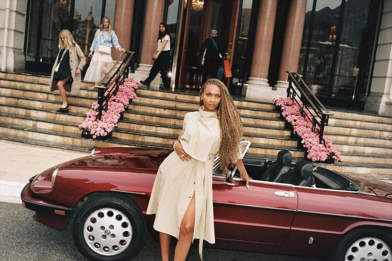 Woman in tan dress poses next to a red convertible car. Other women walk on steps of building with pink flowers in background.