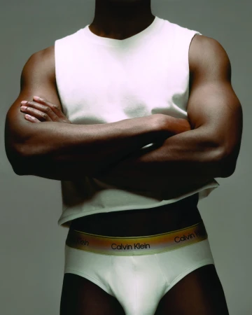 Cropped studio portrait of a muscular Black man in a white sleeveless top and Calvin Klein Pride rainbow briefs with arms crossed.