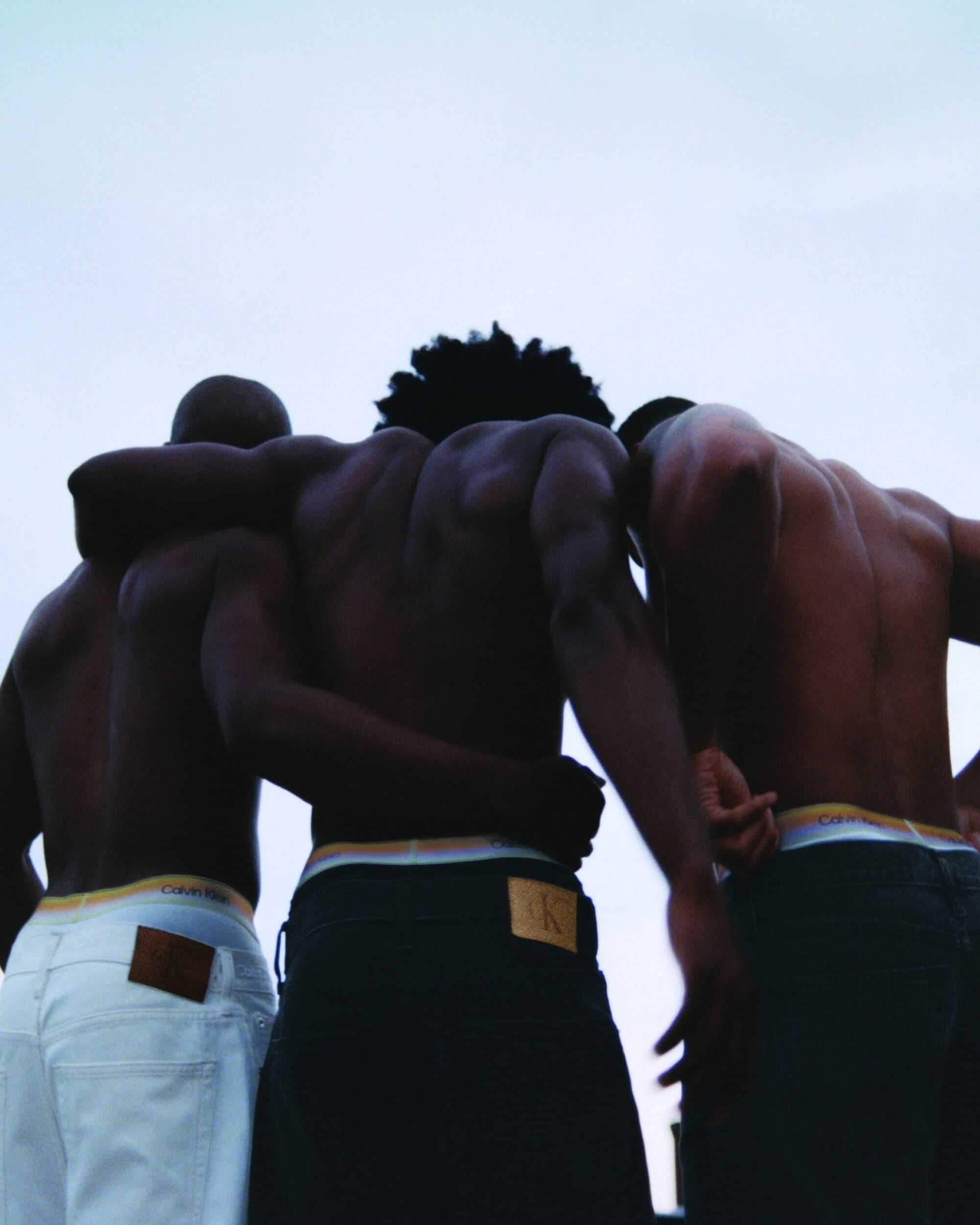 Three shirtless Black men seen from behind embrace with their arms linked against an overcast sky wearing Calvin Klein denim.