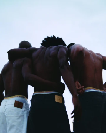 Three shirtless Black men seen from behind embrace with their arms linked against an overcast sky wearing Calvin Klein denim.