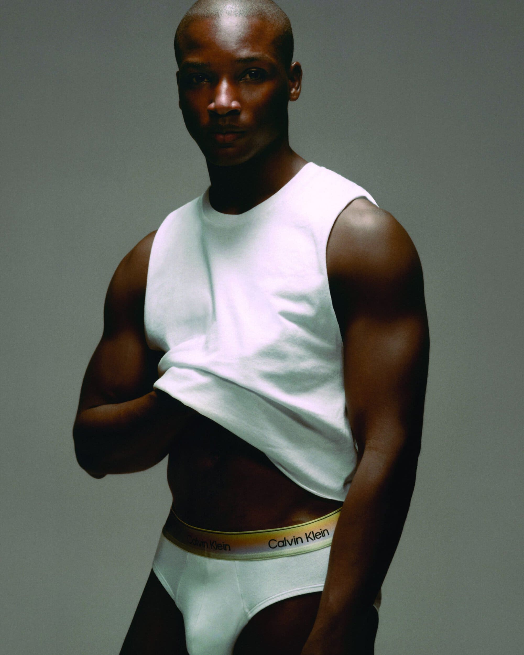 Black man in a white tank top and white briefs against a solid grey background