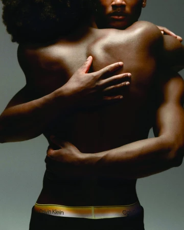 Two Black men embracing closely in a studio setting, one wearing black Calvin Klein underwear with a gradient waistband.