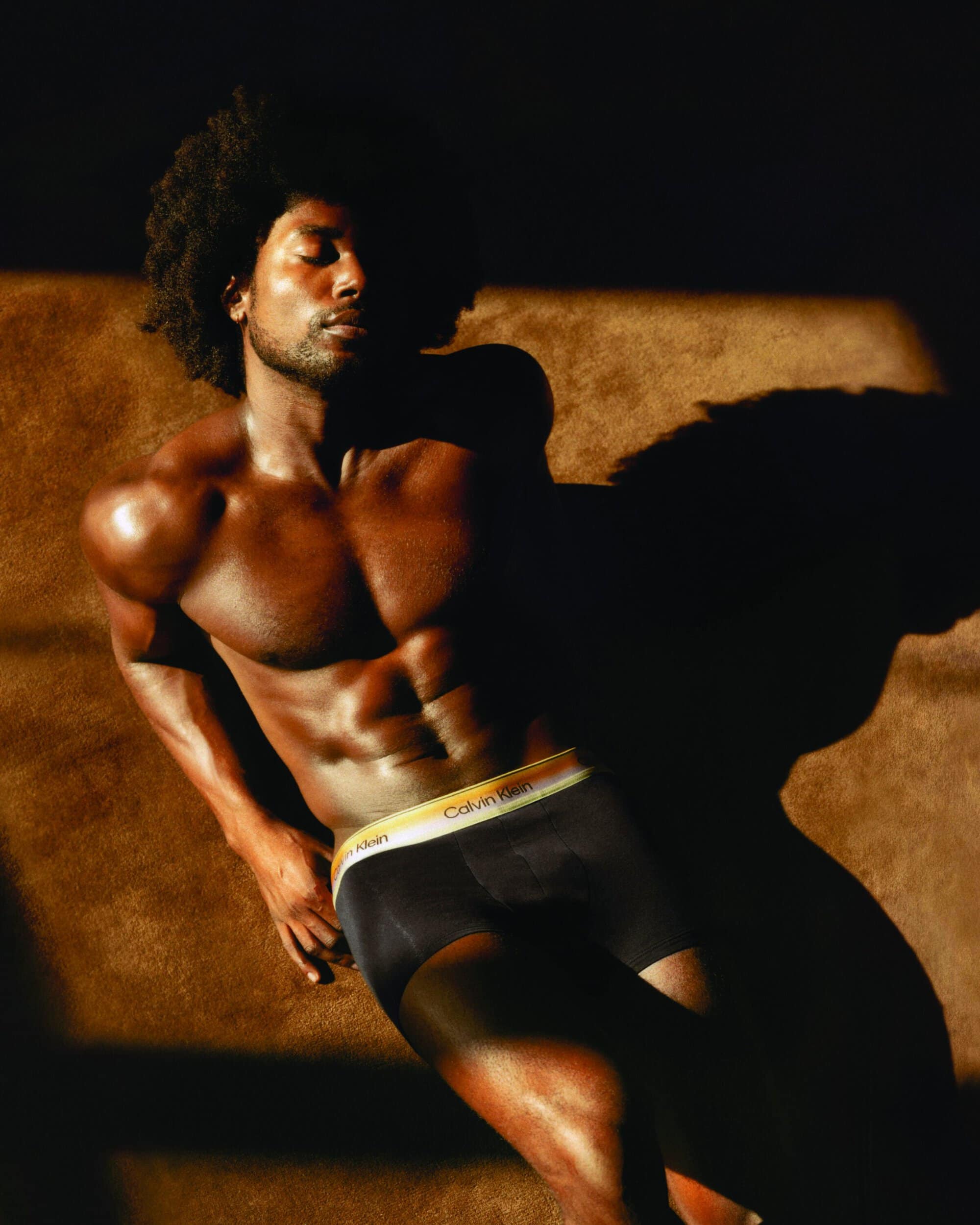Shirtless Black man with an afro lying down in golden sunlight wearing black Calvin Klein boxer briefs with a yellow waistband.
