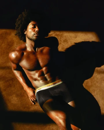 Shirtless Black man with an afro lying down in golden sunlight wearing black Calvin Klein boxer briefs with a yellow waistband.