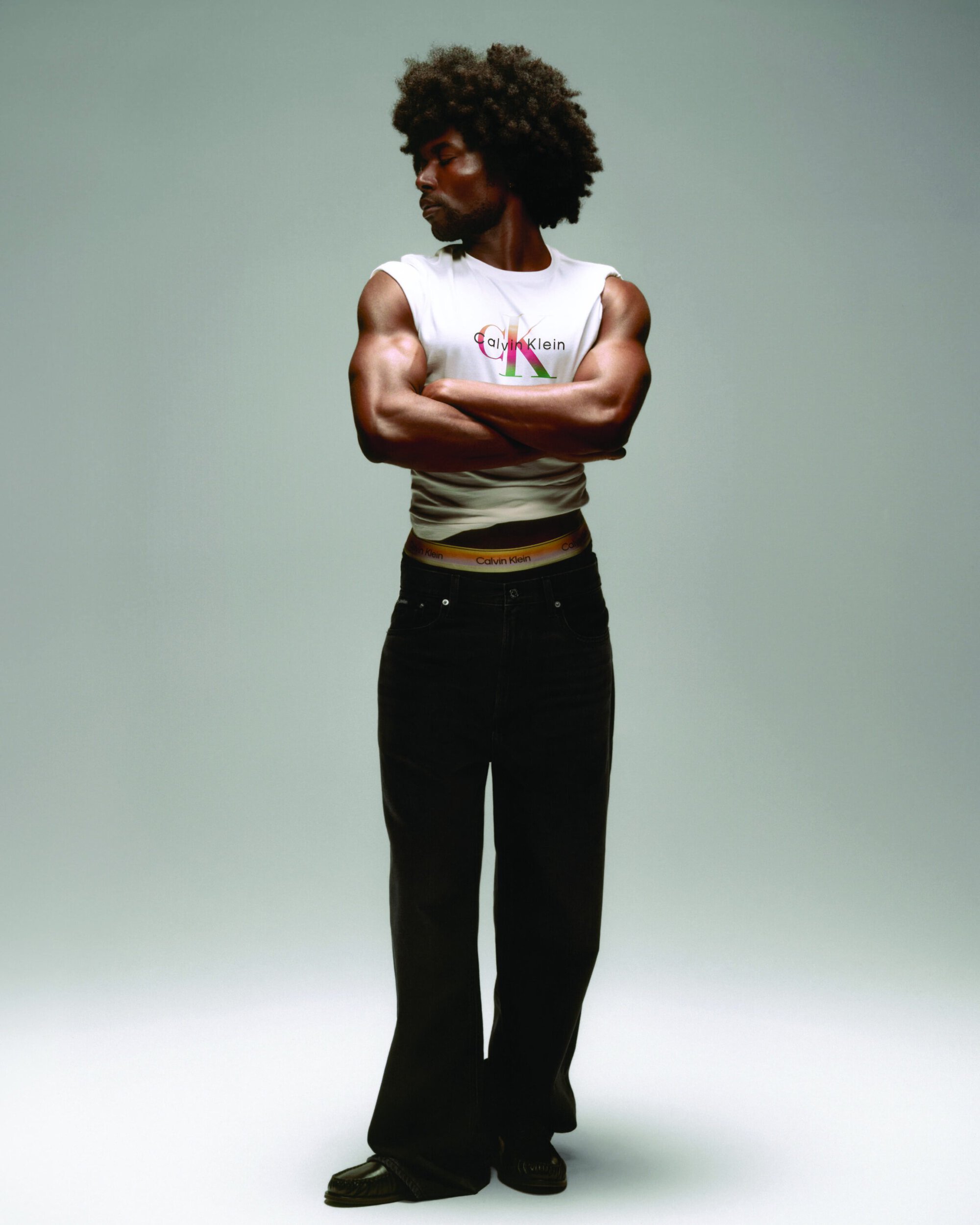 Black male model with an afro standing with arms crossed in a white Calvin Klein top and black jeans against a light studio backdrop.