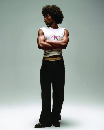 Black male model with an afro standing with arms crossed in a white Calvin Klein top and black jeans against a light studio backdrop.