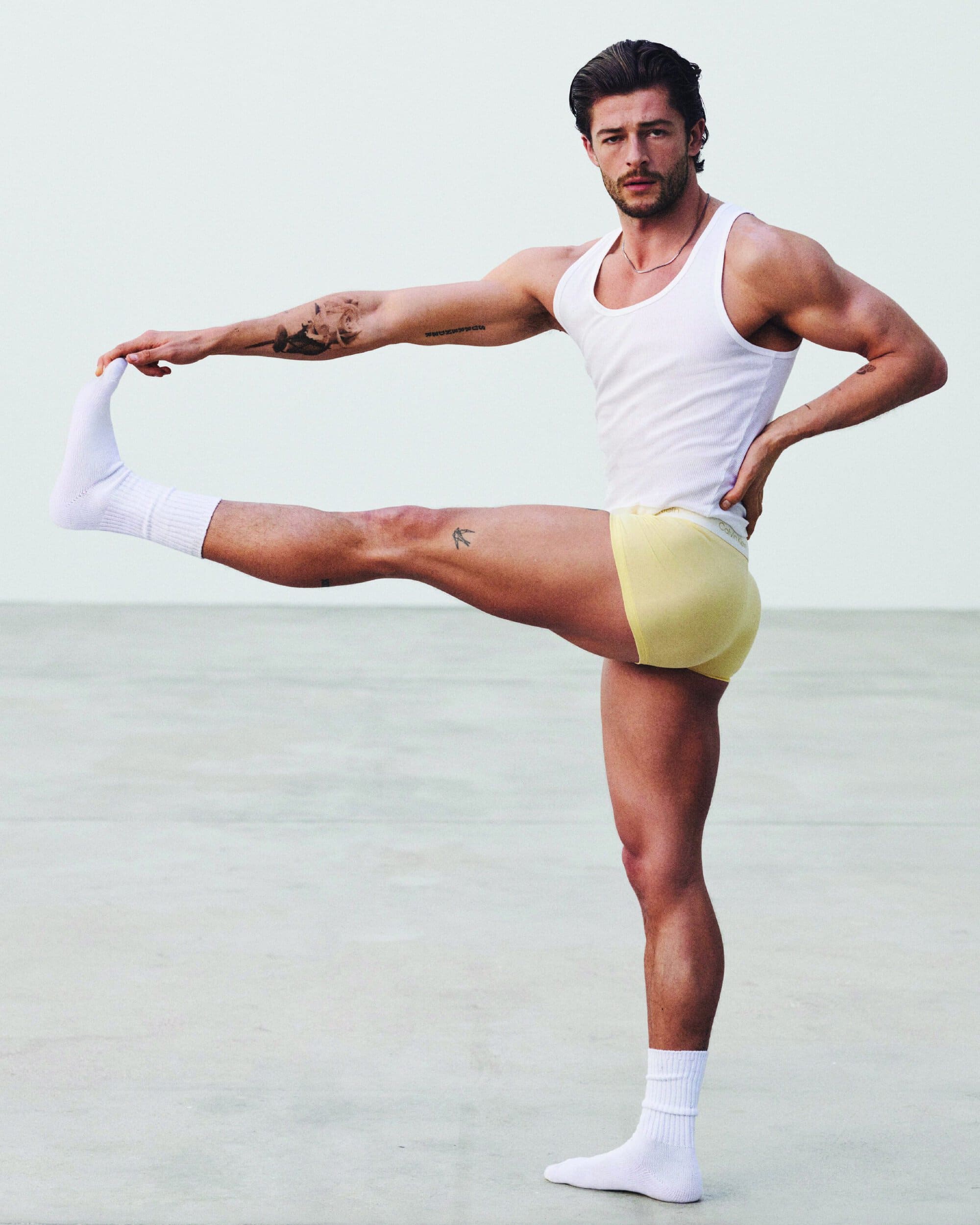 Muscular man wearing a white tank top and yellow briefs stretching his leg against a plain studio background.