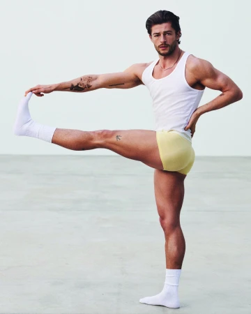 Muscular man wearing a white tank top and yellow briefs stretching his leg against a plain studio background.
