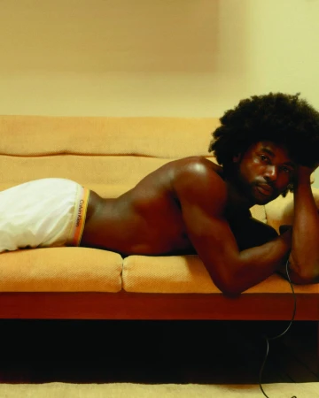 A shirtless Black man with an afro rests on a yellow sofa wearing white Calvin Klein underwear.