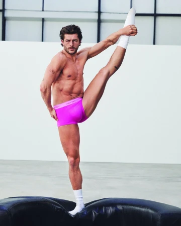 Man in bright pink boxer briefs and white crew socks performs a high vertical leg extension on a black cushion in a bright studio.