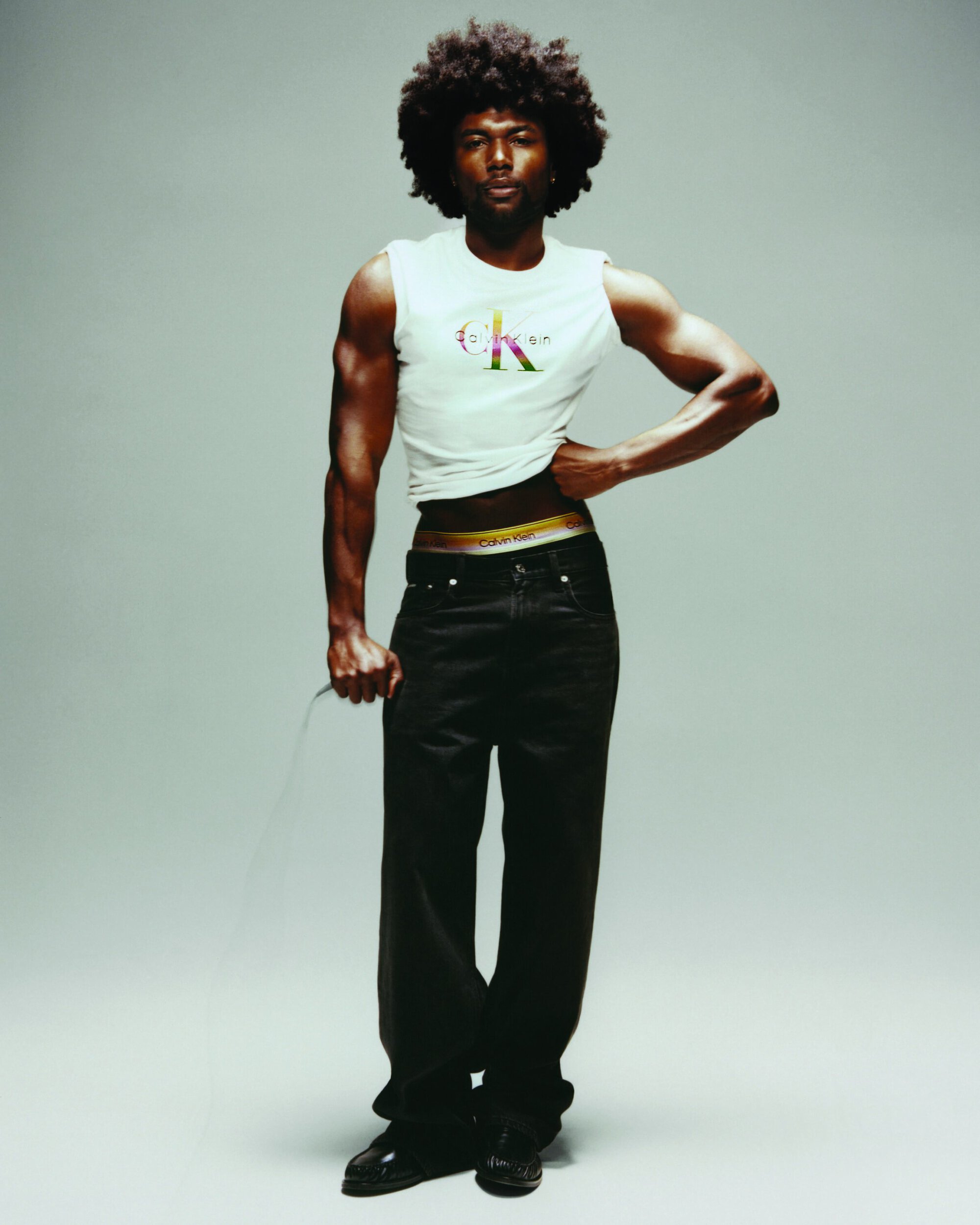 Black man with an afro wearing a white graphic tank top and black jeans against a grey studio backdrop for Calvin Klein.