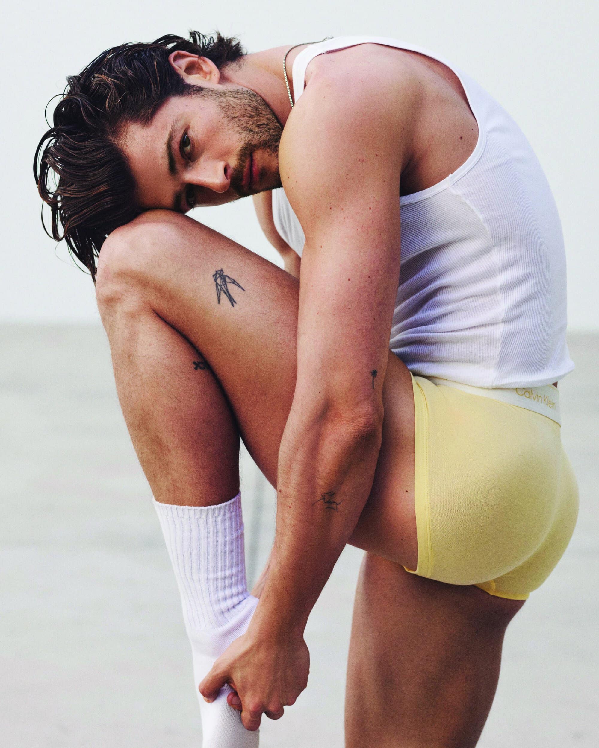 Man in a white ribbed tank top and yellow boxer briefs crouched against a bright white studio backdrop.