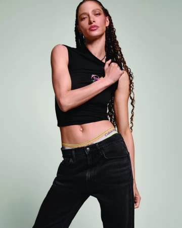Model with long braided hair wearing a black sleeveless top and black jeans against a pale green background