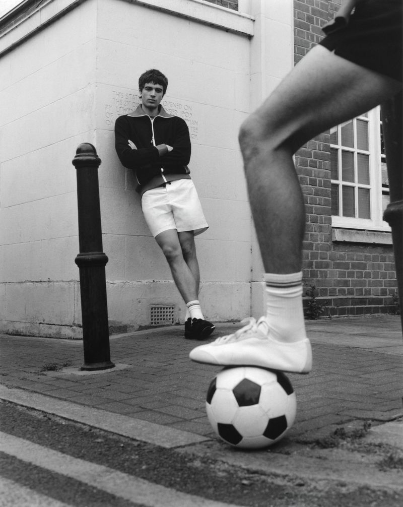A man in a track jacket and shorts leaning against a wall behind a person with their foot resting on a soccer ball.