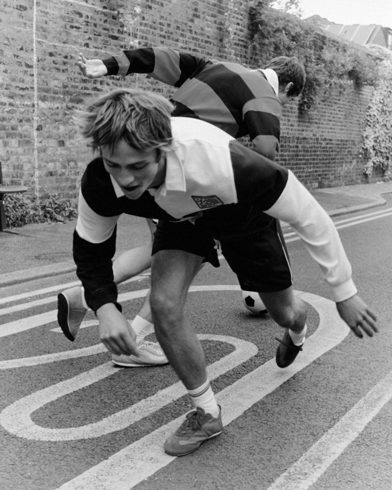 Black and white photo of a young man lunging forward while playing street soccer in a rugby-style jersey and athletic shorts.