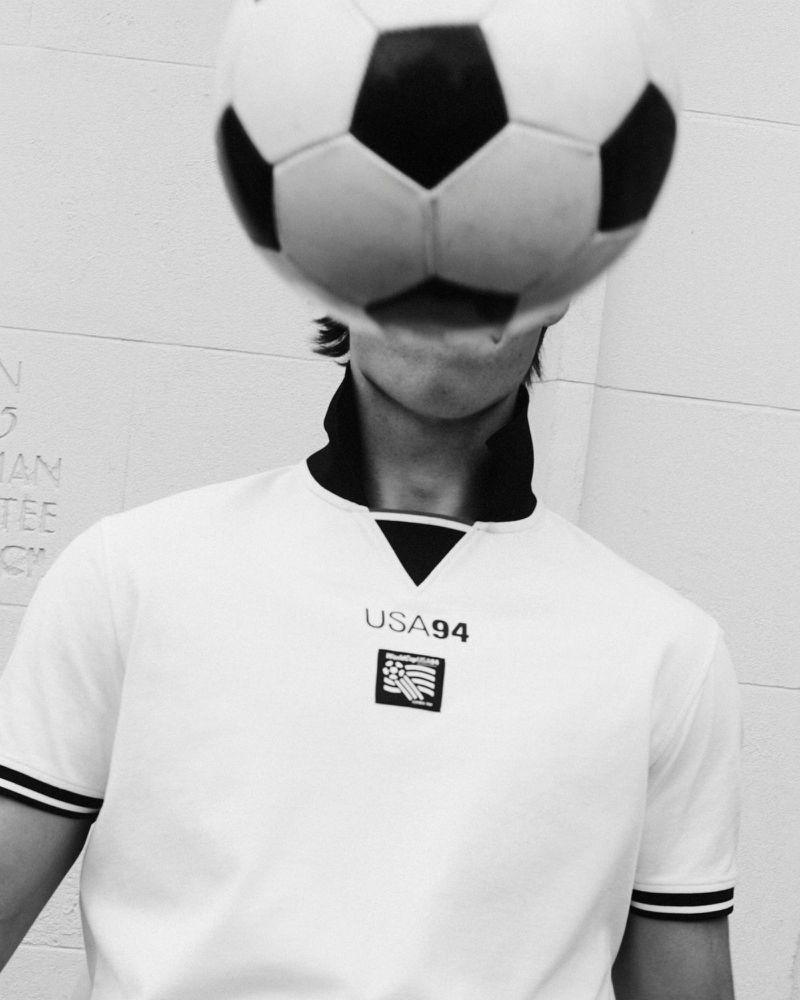 Model wearing a white USA 94 soccer jersey with a black and white soccer ball balancing over their face against a pale wall.