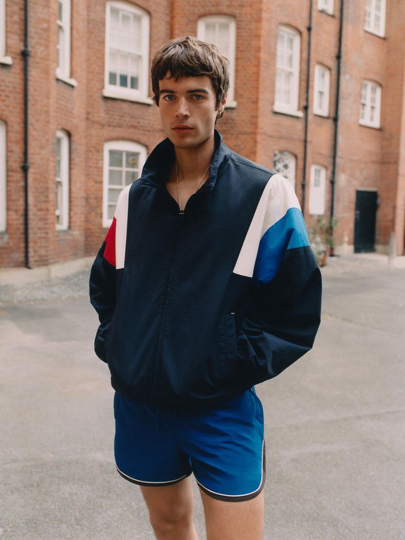 Young man posing in a colorblock windbreaker jacket and blue athletic shorts outside a red brick apartment building.