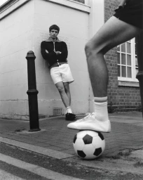 A man in a track jacket and shorts leaning against a wall behind a person with their foot resting on a soccer ball.