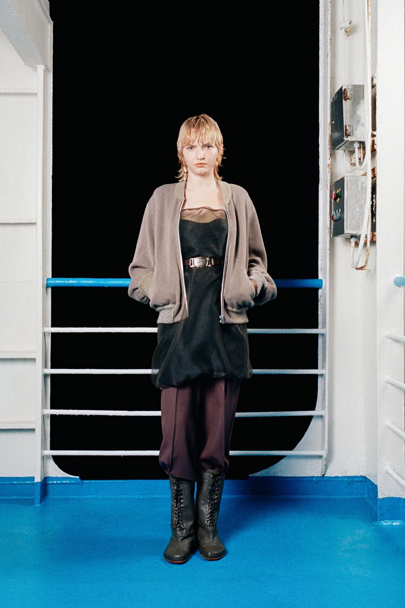A fair-skinned young adult woman stands on the deck of a ship wearing a cardigan, dress with a belt, high-waisted trousers, and lace-up boots.