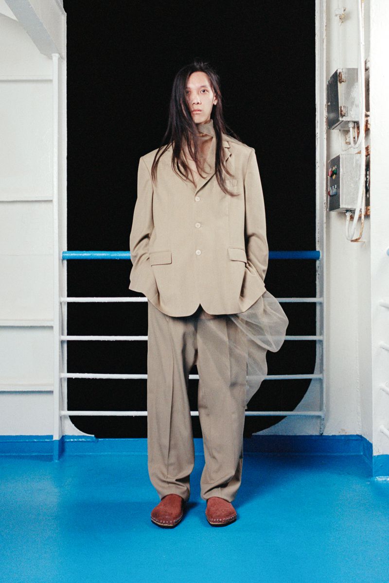 An adult male stands in front of a metal railing wearing a beige suit, tan turtleneck, and brown shoes on a boat, looking at the viewer.
