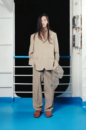 An adult male stands in front of a metal railing wearing a beige suit, tan turtleneck, and brown shoes on a boat, looking at the viewer.