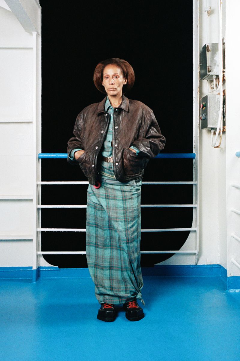 A woman standing in front of a dark background, wearing a brown leather jacket and a plaid skirt on a boat.