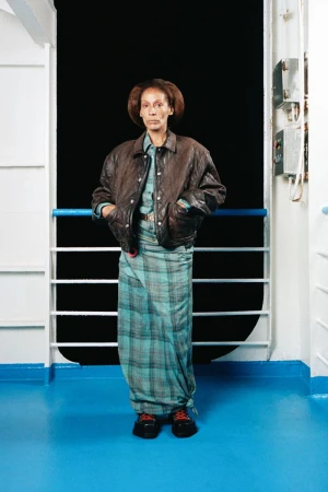 A woman standing in front of a dark background, wearing a brown leather jacket and a plaid skirt on a boat.