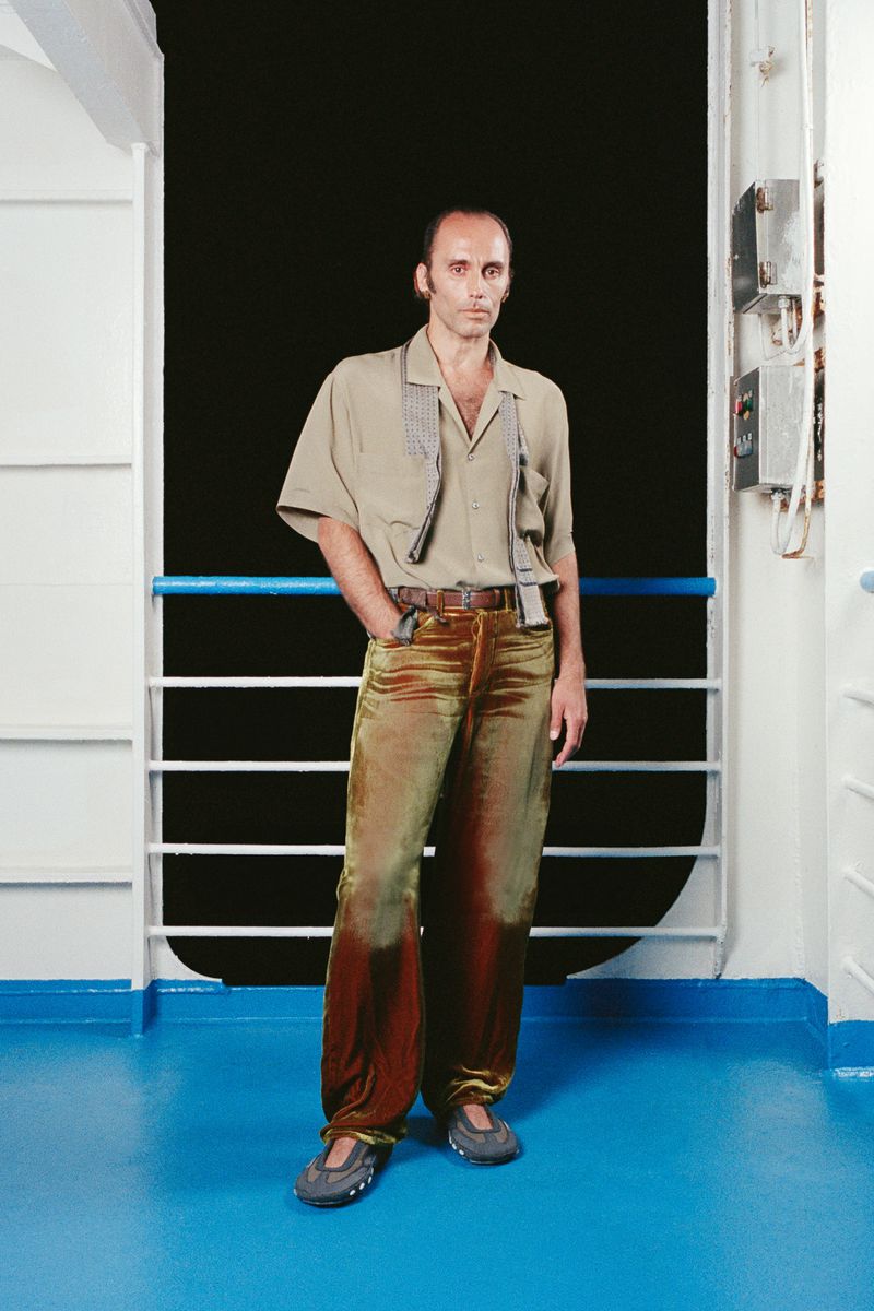 A man in a beige shirt and olive-toned velvet pants stands against a ship railing and window, with a serious facial expression.