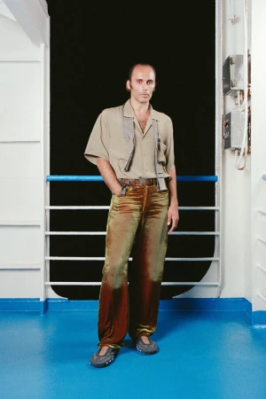 A man in a beige shirt and olive-toned velvet pants stands against a ship railing and window, with a serious facial expression.