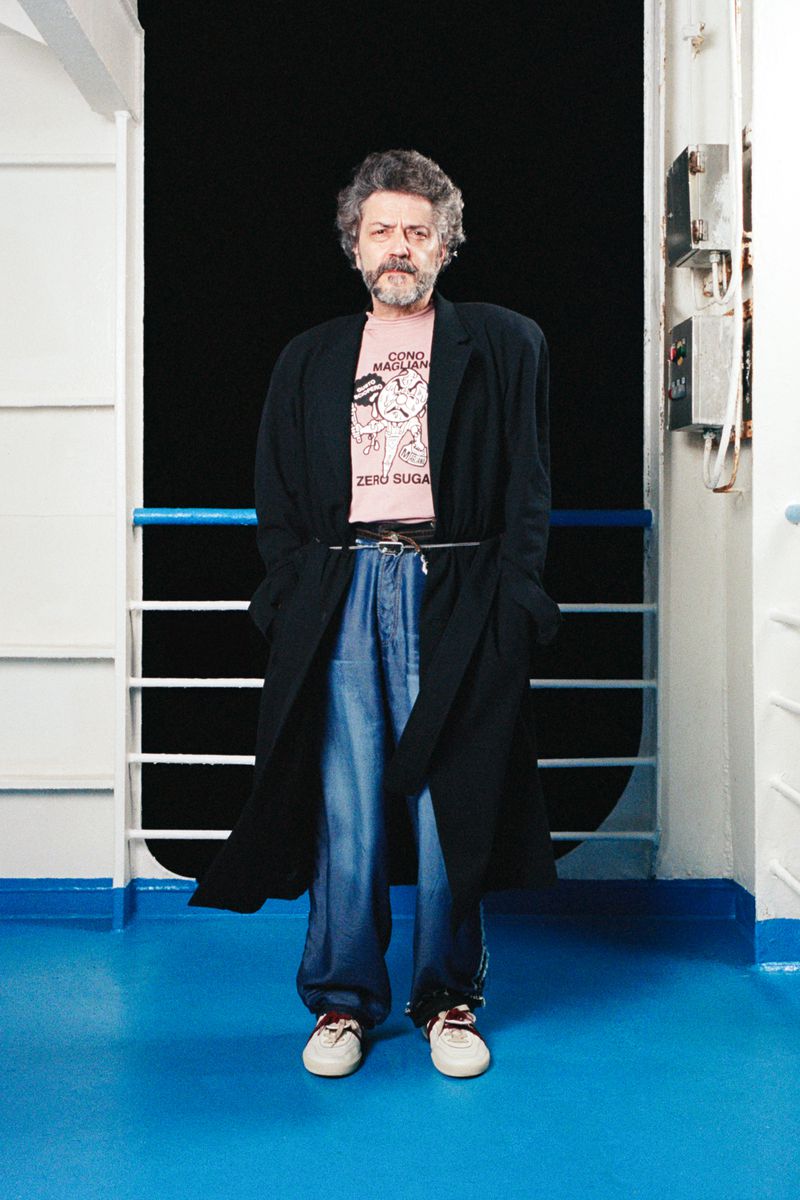 A man stands wearing a black overcoat, jeans, t-shirt, and sneakers on a ship, with a black background behind him, and a blue floor.