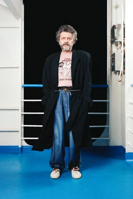 A man stands wearing a black overcoat, jeans, t-shirt, and sneakers on a ship, with a black background behind him, and a blue floor.