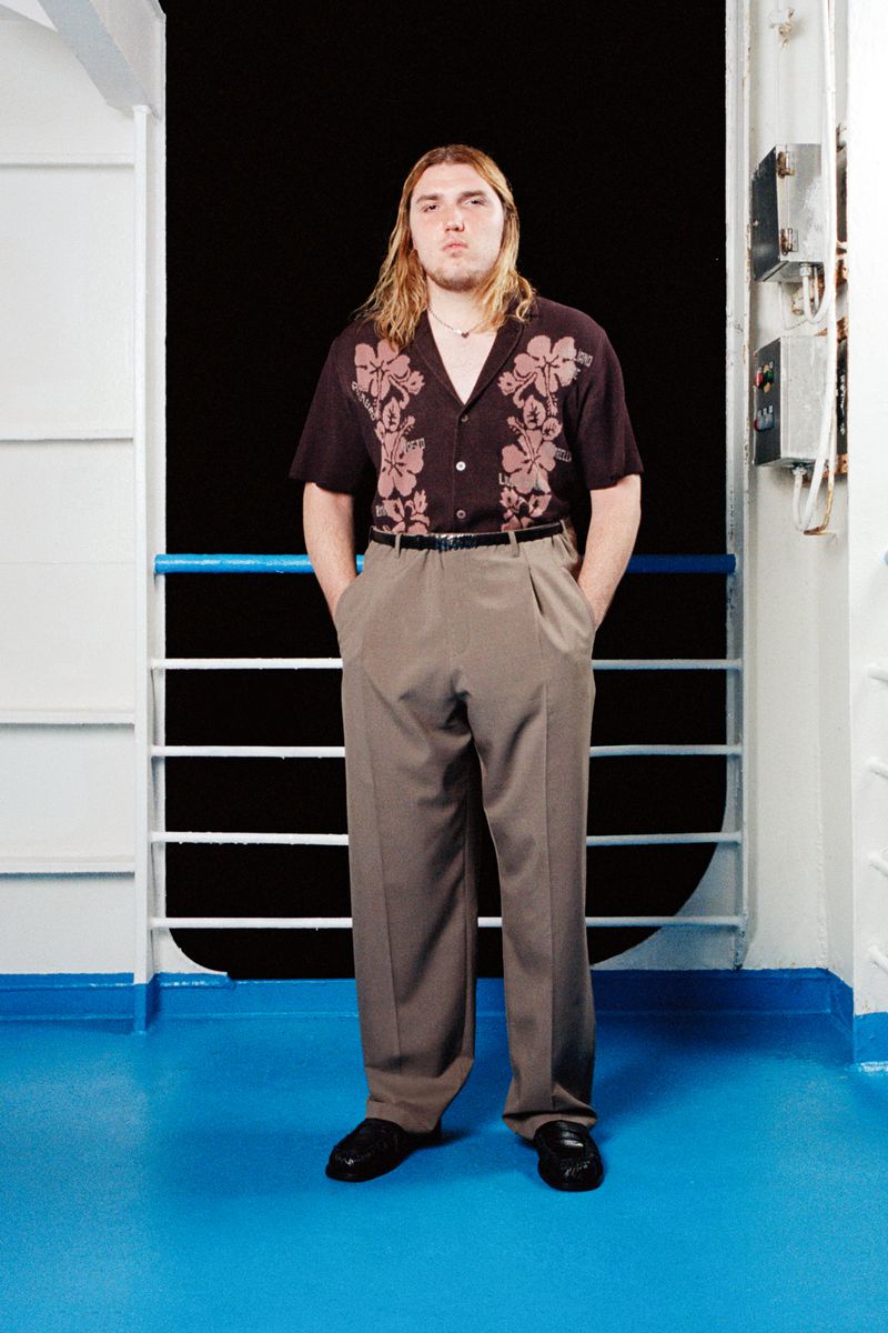 Full-length portrait of a man with long blonde hair in a floral shirt and dress pants posing on a blue and white ship deck.