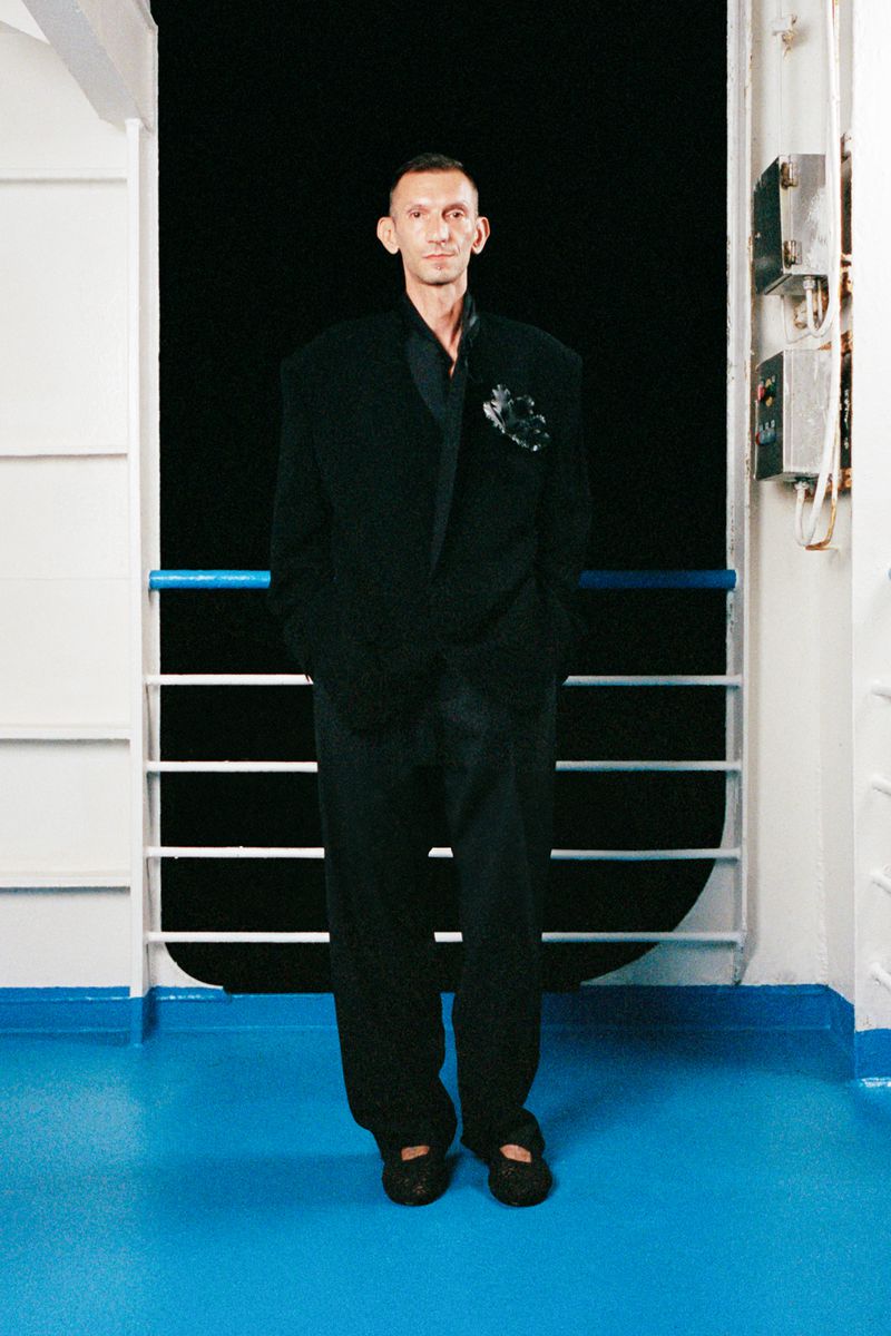 A man in a black suit stands on a blue floor against a dark background. He is framed in a full shot with neutral expression.