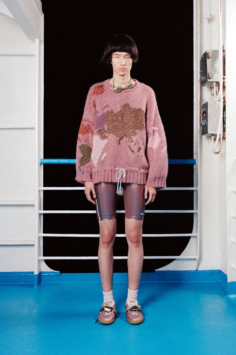 Studio shot of Asian male model wearing a pink sweater and shiny purple shorts standing in front of a white metal wall with black background.