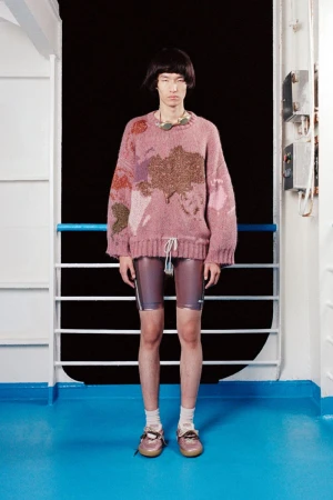 Studio shot of Asian male model wearing a pink sweater and shiny purple shorts standing in front of a white metal wall with black background.
