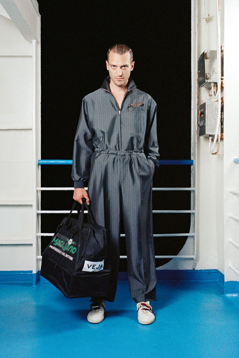Man in pinstripe jumpsuit on a ship deck holds a black sports bag with logos, posing on the blue floor with a black background.