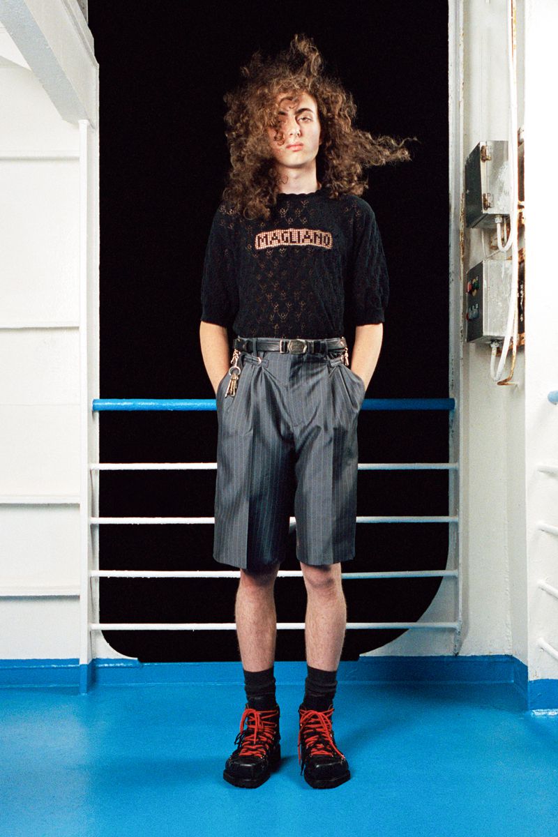 Young adult stands on a blue boat deck wearing a black MAGLIANO shirt, grey pinstripe shorts, black socks, and red-accented hiking boots.